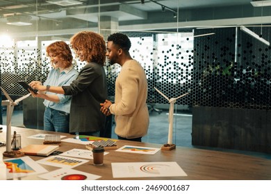 Smiling multicultural environmentalists standing at conference room with tablet in hands and working on renewable energy sources development. Diverse eco staff doing statistics for alternative energy. - Powered by Shutterstock