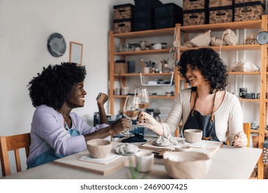 Smiling multicultural pottery course attendees sitting at pottery workshop with wineglasses in hands and making a toast while looking at each other. Diverse pottery class students enjoying wine. - Powered by Shutterstock