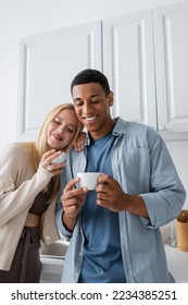 smiling multiethnic couple holding morning coffee in kitchen - Shutterstock ID 2234385251