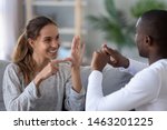 Smiling multiethnic couple or interracial friends talking with sign finger hand language happy two deaf and mute hearing impaired people communicating at home sit on sofa showing hand gestures