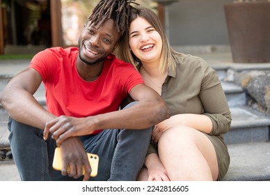 A smiling multi-ethnic couple sitting outside looking at the camera smiling  - Powered by Shutterstock