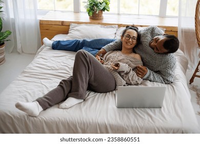 Smiling multiethnic couple in warm sweaters talking near laptop on bed at home - Powered by Shutterstock