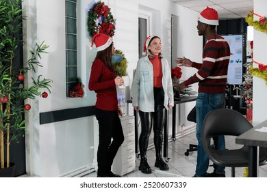 Smiling multiethnic coworkers socialising by office water cooler, discussing Christmas holiday season plans. Relaxed colleagues enjoying conversation during coffee break in festive ornate workspace - Powered by Shutterstock