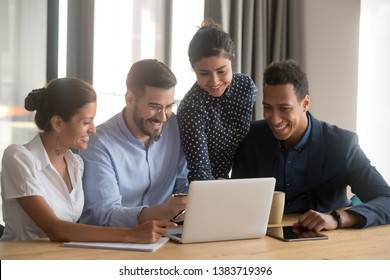 Smiling Multiethnic Employees Sit At Office Desk Look At Laptop Screen Cooperating In Office, Happy Diverse Colleagues Laugh Watching Funny Video On Computer, Brainstorm Together At Meeting