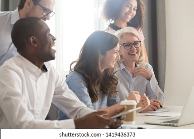 Smiling Multiethnic Employees Sit At Office Desk Laughing Watching Funny Video Training On Laptop, Happy Diverse Workers Or Businesspeople Work Together In Office Have Fun During Coffee Break