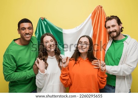 Smiling multiethnic men holding Irish flag near friends isolated on yellow 