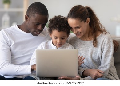 Smiling multiethnic parents relax at home with cute little biracial son using laptop together, happy international family with small boy child sit in couch watch video or play game on computer