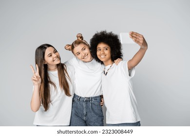 Smiling multiethnic teen friends in white t-shirts taking selfie on smartphone, hugging and gesturing while standing isolated on grey, teenagers bonding over common interest, friendship - Powered by Shutterstock