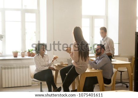 Smiling multiracial colleagues talking eating pizza at lunch in office room, diverse team people enjoy meal at coffee break chatting together, food delivery or good friendly relations at work concept
