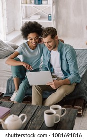 Smiling Multiracial Couple Looking At Laptop Screen While Sitting On Bed