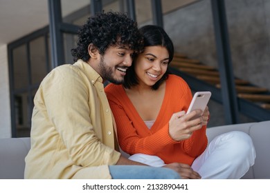 Smiling Multiracial Friends Using Mobile Phone Shopping Online Relaxing At Home. Happy Indian Man And African American Woman Playing Mobile Game Looking At Digital Screen 