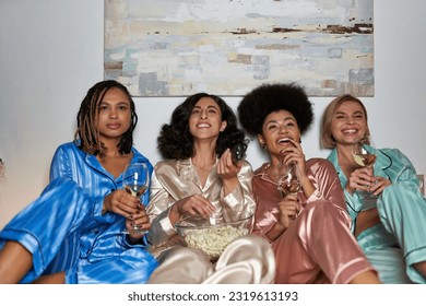 Smiling multiracial women in pajama watching tv and holding popcorn and glasses of wine while sitting on bed during girls night at home, bonding time in comfortable sleepwear, slumber party