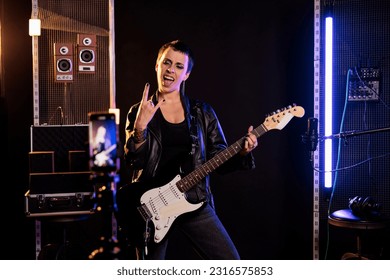 Smiling musician doing rock sign while playing at electric guitar in front of smartphone recording heavy metal song, preparing new rock album. Rebel guitarist performing grunge music in sound studio - Powered by Shutterstock