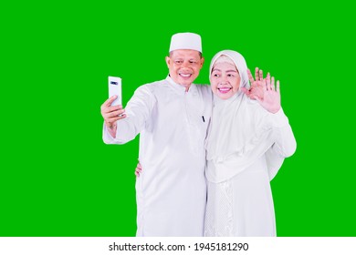 Smiling Muslim Aged Couple Doing Video Call With Their Family On A Mobile Phone During Eid Mubarak While Standing In The Studio With Green Screen Background