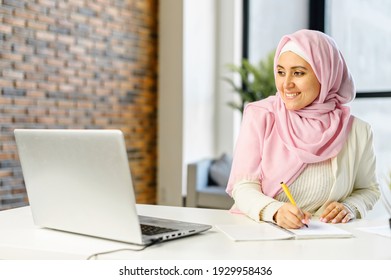 Smiling muslim businesswoman taking notes sitting at the desk in modern office space. Young islamic female student wearing hijab and smart casual wear overwrites from the laptop screen into notebook - Powered by Shutterstock