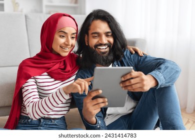 Smiling muslim couple with digital tablet relaxing on floor at home, happy arab man and woman in hijab sharing fun moment while looking at tablet, browsing internet or watching videos, closeup - Powered by Shutterstock