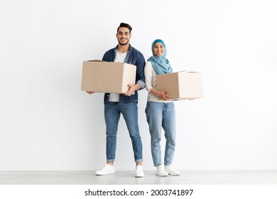 Smiling muslim couple moving to new apartment, standing together over white wall with big paper boxes full of their belongings, copy space, full length shot. Moving, relocation, real estate concept