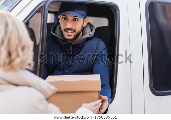 smiling muslim delivery man giving parcel to\
client on blurred\
foreground
