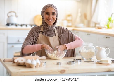 Smiling Muslim Lady In Hijab Baking In Kitchen, Kneading Dough And Smiling At Camera. Cheerful Islamic Woman Wearing Apron, Having Fun At Home And Enjoying Cooking Homemade Food, Free Space - Powered by Shutterstock