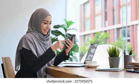 Smiling Muslim Lady Texting On Mobile Phone While Working On Computer Laptop In Office