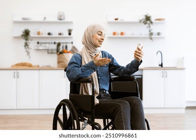 Smiling Muslim woman in wheelchair using smartphone in modern home kitchen. Concept of disability, technology, and independence. Wheelchair user wearing hijab engaging in video call. - Powered by Shutterstock