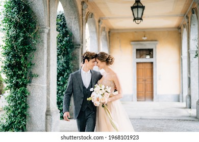 Smiling newlyweds embrace while walking along the terrace with columns against the backdrop of greenery. Lake Como