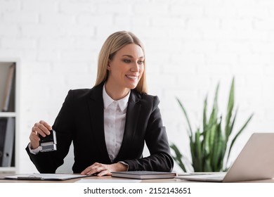 Smiling Notary In Formal Wear Holding Stamper And Looking At Laptop In Office