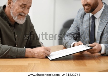 Smiling notary showing senior man where to sign Last Will and Testament at wooden table indoors, closeup