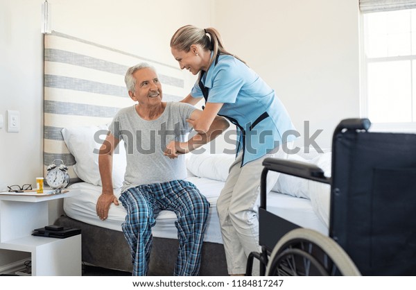 Smiling nurse assisting senior man to get up from\
bed. Caring nurse supporting patient while getting up from bed and\
move towards wheelchair at home. Helping elderly disabled man\
standing up.