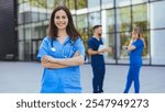 Smiling nurse in blue scrubs stands confidently outside a modern building, with two colleagues engaged in conversation in the background. Represents teamwork and professionalism in the healthcare 