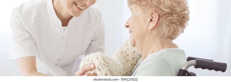 Smiling Nurse Lying A Warm Blanket On Her Elderly Patient Sitting In A Wheelchair In A Daycare Center