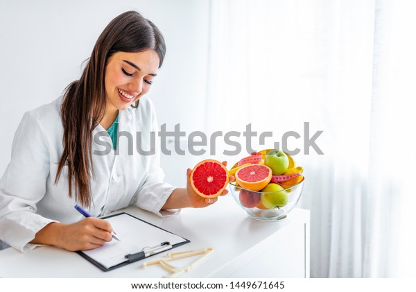 Smiling nutritionist in her\
office, she is showing healthy vegetables and fruits, healthcare\
and diet concept. Female nutritionist with fruits working at her\
desk. 