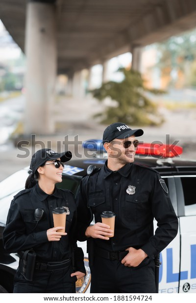 Smiling officers of\
police in sunglasses holding coffee to go near auto on blurred\
background on urban\
street