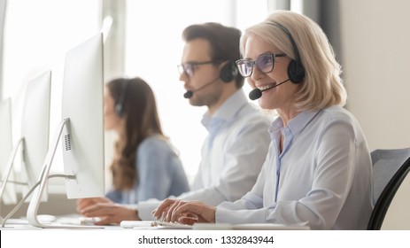 Smiling Old Female Call Center Agent Operator Telemarketer In Headset Talking Consulting Online Client At Workplace, Middle Aged Businesswoman Using Computer Works In Customer Service Support Office