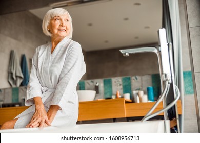 Smiling Old Lady In Bathrobe Sitting On The Edge Of Bathtub Stock Photo