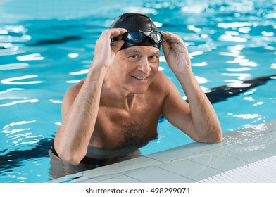 Smiling Old Man In A Swimming Pool Wearing Swim Cap And Goggles On His Head. Portrait Of A Satisfied Senior Man After Swimming In The Pool. Retired Proud Man Leaning At The Poolside Looking At Camera.