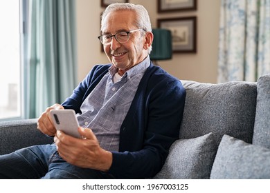 Smiling old man using smartphone while sitting on couch. Happy elderly man with spectacles messaging on mobile phone at home. Senior in video call with his family on smart phone at nursing home.