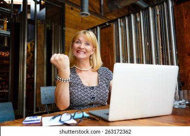 Smiling Old Senior Blonde Business Woman 60-65 Years, Alone With Laptop In Summer Cafe With Gesture On Face And Raised Hand Up With Fist