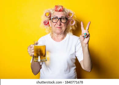 Smiling Old Woman With Curlers With A Glass Of Beer Shows A Gesture Of Two Fingers, Victories And Peace On A Yellow Background