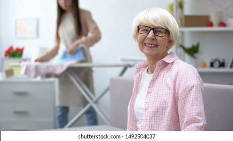 Smiling Old Woman Looking At Camera, Granddaughter Ironing Clothes On Background