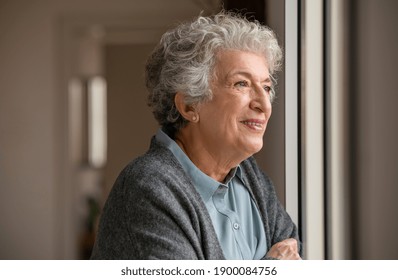 Smiling old woman looking through the window. Happy senior woman standing near window during quarantine due to covid. Retired and contemplative lady wearing sweater while looking outside and thinking.