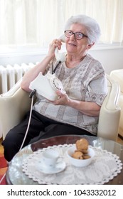 Smiling Old Woman Talking On Landline Rotary Phone While Relaxing On Armchair At Home.