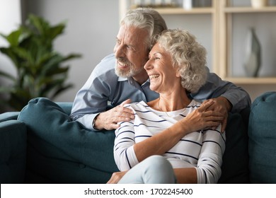 Smiling older couple dreaming about good future together, looking out window, loving aged husband hugging wife, sitting on cozy couch at home, happy mature man and woman lost in thoughts