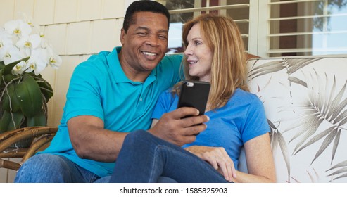 Smiling Older Couple Sitting On Porch And Using Cellphone Together. African American And Caucasian Husband And Wife Looking At Mobile Phone On Sunny Day Outside Home