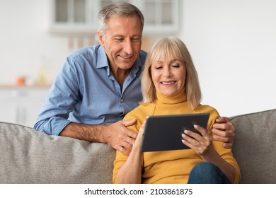 Smiling Older Couple Using Tablet Computer Watching Movie Online Together And Browsing Internet Sitting On Sofa At Home On Weekend. Mature Couple Using Digital Gadget. Technology Concept