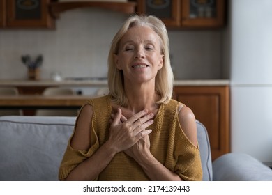 Smiling Older Female Sincere Put Folded Arms On Chest Praying Or Say Thank You To God For Help, Luck Or Life Miracle Seated On Sofa With Eyes Closed At Home. Faith, Religion, Express Gratitude Concept