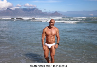 Smiling older fitness man in white speedo standing in water with Table Mountain in background
