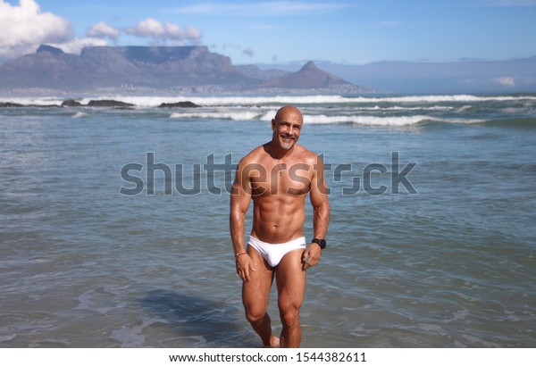 Smiling older fitness man in white\
speedo standing in water with Table Mountain in\
background