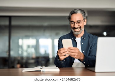 Smiling older Indian business man sitting at desk using mobile phone. Happy mature businessman executive, busy middle aged entrepreneur investor looking at smartphone working in office with cellphone. - Powered by Shutterstock