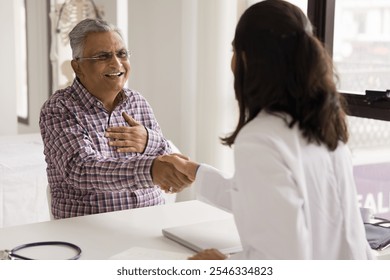 Smiling older Indian male patient sit at table in clinic during visit giving handshake to young female general practitioner, express respect, gratitude for got professional medical care or retirement - Powered by Shutterstock